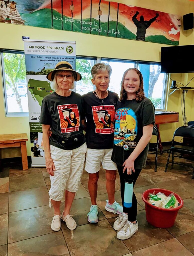 Three women standing and smiling with dark t-shirts. They are in a classroom.