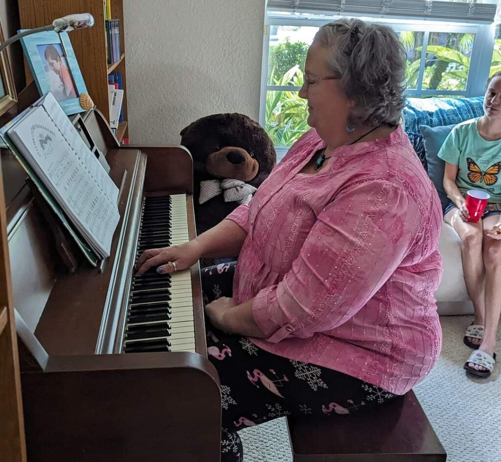 A lady sits and plays a piano - she is looking at the piano and has a pink shirt on.