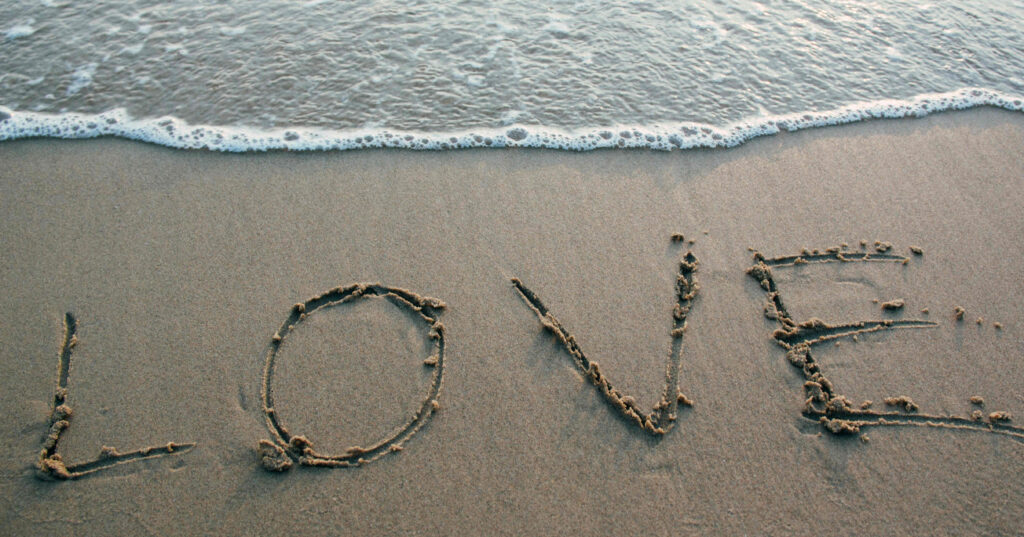 A dark tan image of sand with water at the top and the word 'love' written in the sand